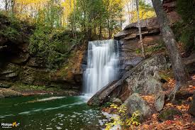 A rugged 110-foot waterfall system with three cascading tiers, nestled in a forested gorge near Lake Superior, featuring moss-covered rocks and wildflower trails.