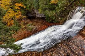 A towering 100-foot waterfall cascading over sandstone cliffs in Michigan’s Upper Peninsula, surrounded by dense forest and accessed via a wooden staircase.