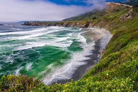 Crescent-shaped sandy cove with tidepools, driftwood logs, and cliffs dotted with Monterey cypress trees.