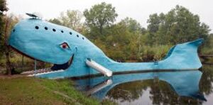 A smiling bright-blue whale statue in a pond, featuring a slide on its tail and picnic tables alongside Route 66