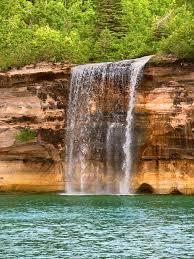 A dramatic 70-foot waterfall plunging directly into Lake Superior at Pictured Rocks, mist swirling into rainbows against layered sandstone cliffs.