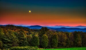 A rural New England landscape of rolling Green Mountains, dotted with white-steepled villages, red barns, maple farms, and covered bridges under golden fall foliage or snowy winter skies.